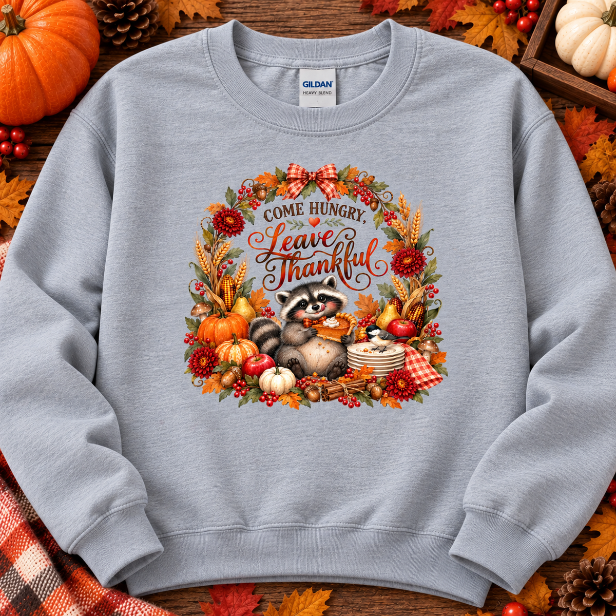A light gray crewneck sweatshirt featuring a colorful autumn-themed graphic of a raccoon surrounded by pumpkins, leaves, and the text 'Come Hungry. Leave Thankful.