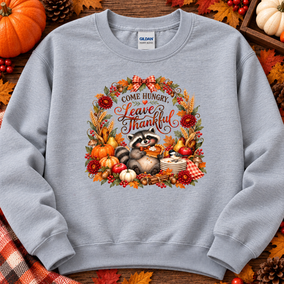 A light gray crewneck sweatshirt featuring a colorful autumn-themed graphic of a raccoon surrounded by pumpkins, leaves, and the text 'Come Hungry. Leave Thankful.