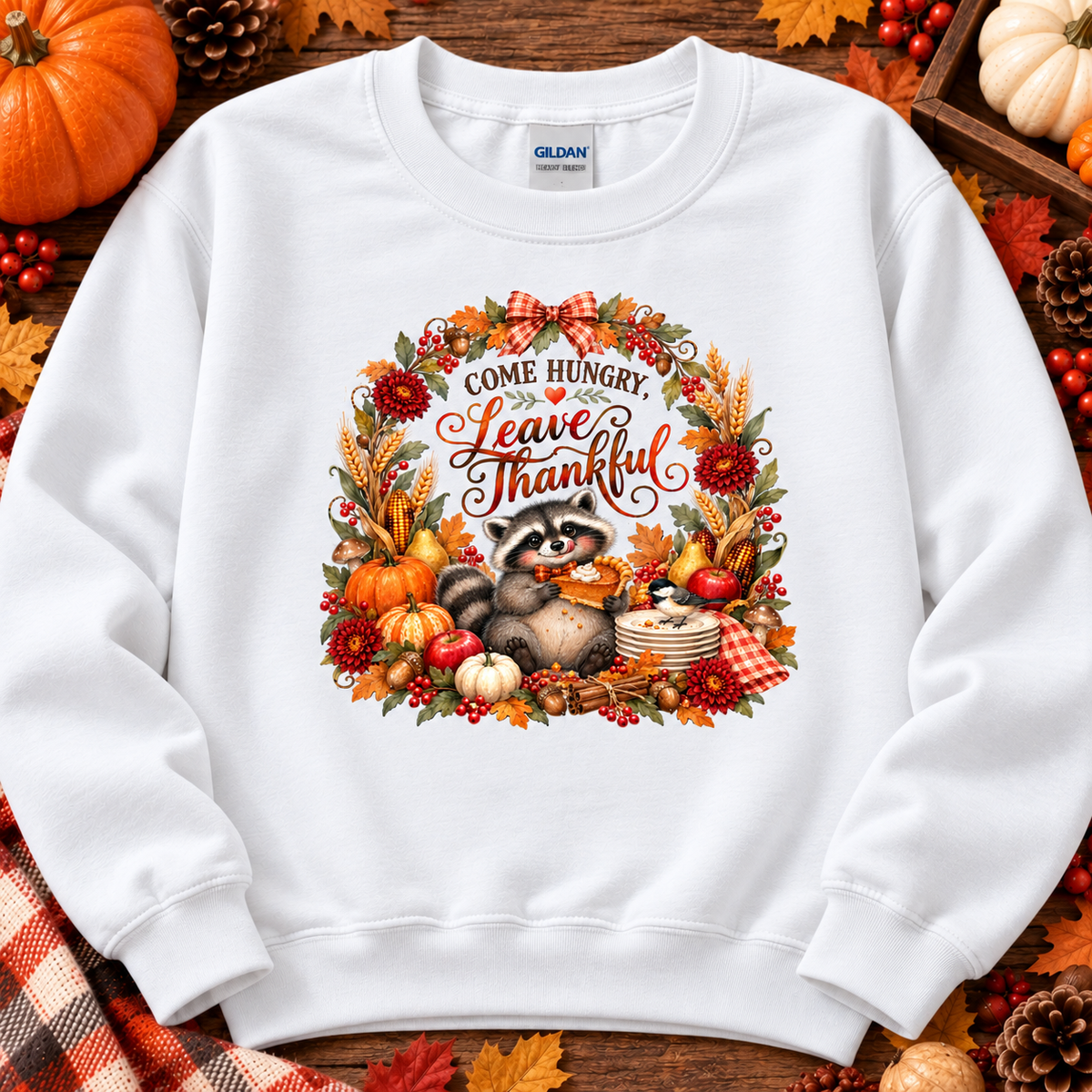 A white Gildan sweatshirt laid flat on a rustic wooden surface surrounded by autumn decorations. The sweatshirt features a colorful graphic of a raccoon holding a corn cob, framed by a wreath of pumpkins, fall leaves, berries, and the text 'COME HUNGRY, LEAVE THANKFUL'.