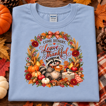 A folded light blue Gildan Softstyle t-shirt with a detailed autumn-themed graphic print. The print features a raccoon holding a plate of food, surrounded by a wreath of pumpkins, corn, berries, and the text 'COME HUNGRY, LEAVE THANKFUL'.