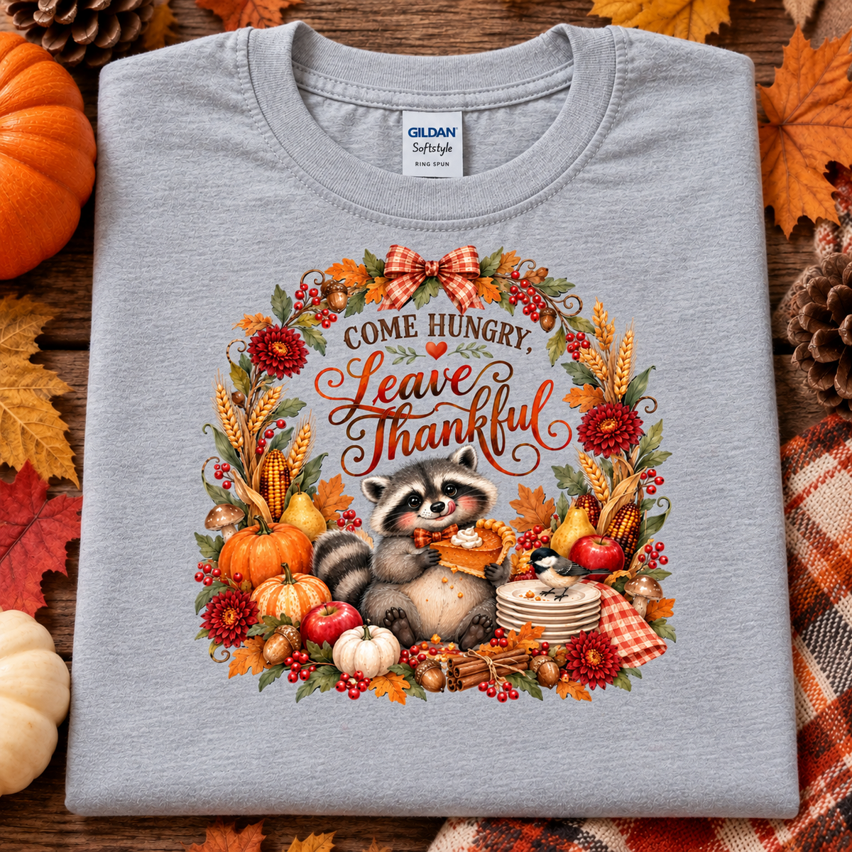 A folded light heather gray Gildan Softstyle t-shirt with a detailed autumn-themed graphic print. The graphic features a raccoon holding a corn cob, surrounded by a wreath of fall foliage, pumpkins, corn, and the text 'COME HUNGRY, Leave Thankful'.