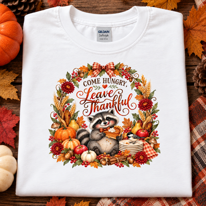 A folded white Gildan Satinway t-shirt with a detailed autumn-themed graphic print. The graphic features a raccoon holding a pumpkin inside a wreath with the text 'COME HUNGRY.