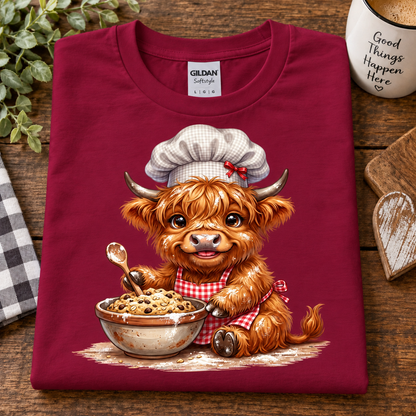 A folded cardinal red t-shirt with a graphic of a cute Highland cow character wearing a chef's hat and apron, sitting in front of a bowl of cereal. The shirt is displayed on a wooden surface with lifestyle props including a mug, a heart-shaped coaster, and a checkered cloth.