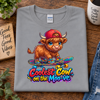 A folded sport grey Gildan Softstyle t-shirt is displayed on a rustic wooden table. The shirt features a colorful graphic of a cute, fluffy Highland cow wearing a red cap and boots, riding a skateboard, with the text 'Coolest Cow on the Moo-ve' written below in a playful multicolor font.