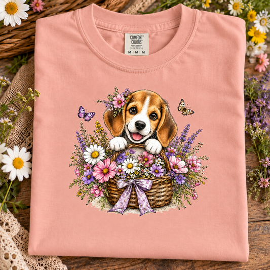 A folded peachy pink t-shirt with a colorful graphic of a beagle puppy in a flower basket, displayed on a rustic wooden surface surrounded by flowers.