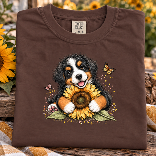 A folded espresso brown Comfort Colors t-shirt featuring a graphic of a Bernese Mountain Dog puppy holding a large yellow sunflower, surrounded by butterflies and small flowers, displayed on a rustic wooden surface with sunflowers in the background.