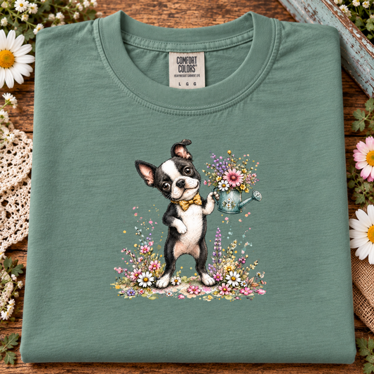 A folded light green t-shirt with a graphic of a Boston Terrier watering a flower garden, displayed on a rustic wooden background with decorative flowers.