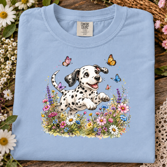 A hydrangea blue colored comfort colors t-shirt with a Dalmatian puppy jumping through a field of colorful wildflowers, surrounded by butterflies, on a plain white background.