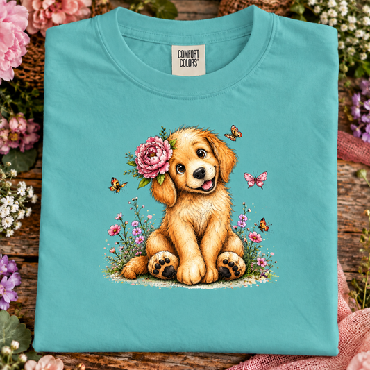 A folded chalky mint Comfort Colors t-shirt displayed on a rustic wooden surface surrounded by flowers. The shirt features a colorful graphic of a golden puppy sitting in a field with a pink rose on its head, surrounded by butterflies and wildflowers.