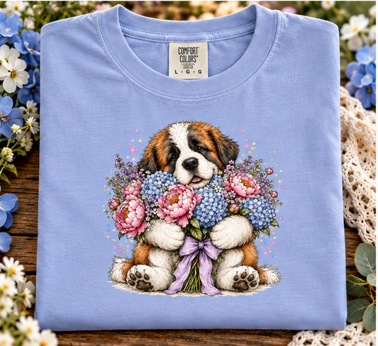 A folded periwinkle light blue Comfort Colors T-shirt with a detailed graphic of a Saint Bernard puppy holding a bouquet of pink and blue flowers, displayed on a rustic wooden surface surrounded by flowers and lace.