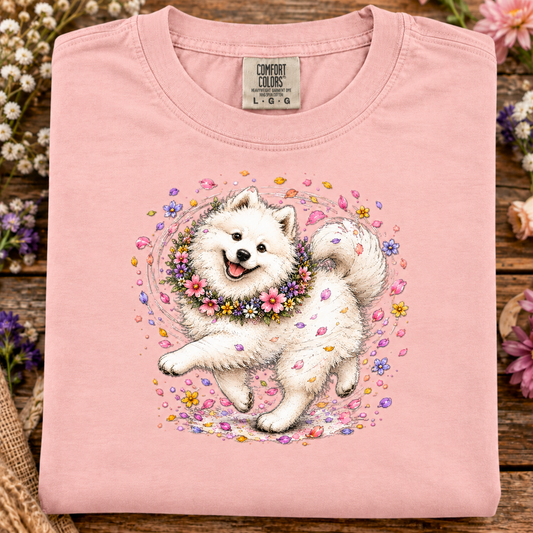 A folded rose quartz pink Comfort Colors t-shirt featuring a colorful graphic of a white Samoyed dog wearing a floral collar, surrounded by pink and yellow flower petals, displayed on a rustic wooden surface with flowers.