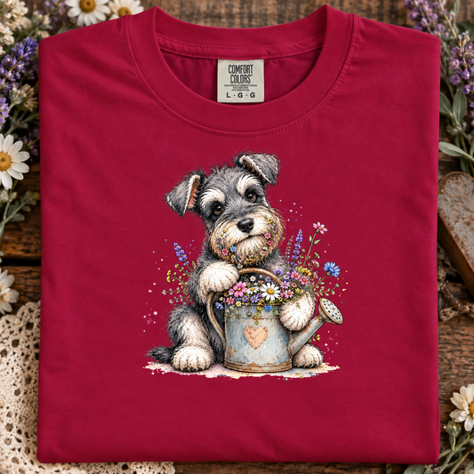 A crimson colored t-shirt with a cute scruffy dog with gray, black, and white fur sits behind a weathered blue watering can filled with colorful wildflowers on a white background.