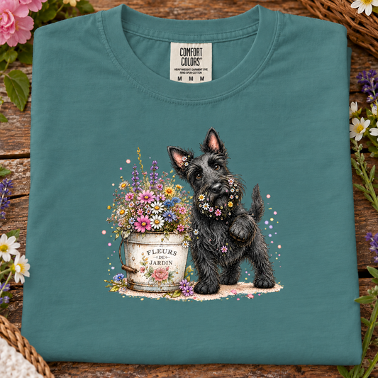 A seafoam folded T-shirt displayed on a rustic wooden surface with flowers and a wicker basket around it. The shirt has a printed illustration of a black scruffy dog standing beside a white flower pot filled with multicolored flowers.