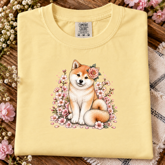 A folded butter yellow Comfort Colors T-shirt with a detailed graphic of a Shiba Inu dog surrounded by pink cherry blossoms, displayed on a rustic wooden surface with decorative flowers.