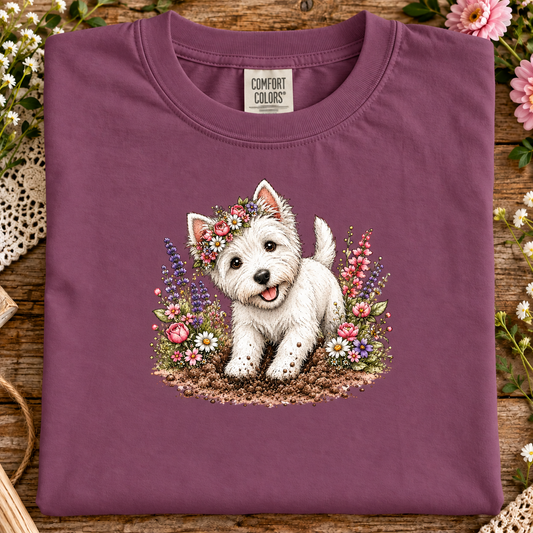 A folded berry purple Comfort Colors t-shirt featuring a detailed graphic of a white West Highland terrier dog surrounded by colorful wildflowers, displayed on a rustic wooden surface with decorative flowers.