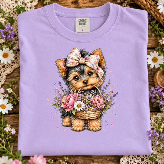 A orchid colored Comfort Colors t-shirt laid flat on a rustic wooden surface, decorated with white and purple flowers. The front of the shirt features a detailed graphic of a cute Yorkshire Terrier puppy wearing a floral bow and holding a basket of colorful flowers.
