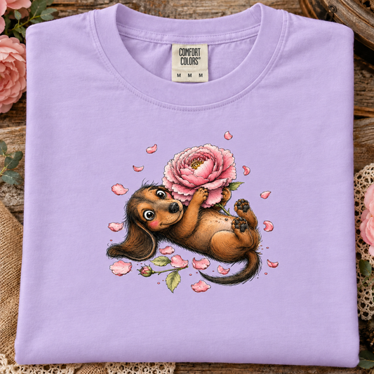 An orchid colored t-shirt with a cute cartoon dachshund lying on its back and holding a large pink peony flower, with scattered pink petals around it on a white background.