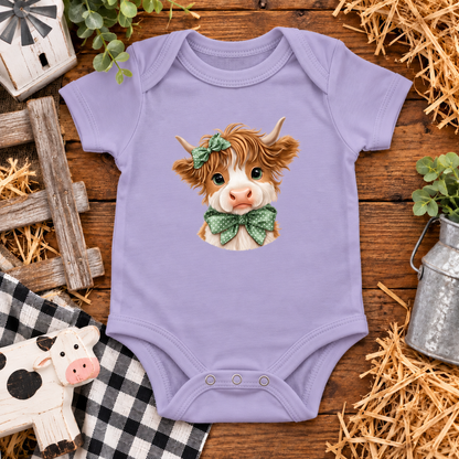 A lavender short-sleeve baby bodysuit laid flat on a rustic wooden surface, featuring a cute illustrated cow face with a green bow tie and bow headband. The bodysuit is styled with farm-themed props including straw, a small wooden fence, a galvanized milk canister, a black-and-white check cloth, and a small cow figurine.