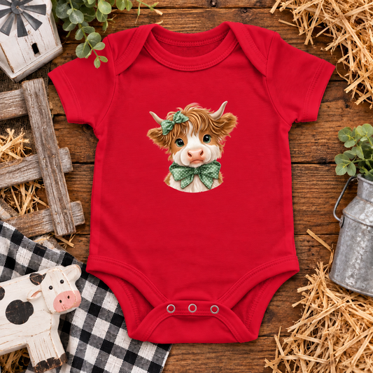 A red short-sleeve baby bodysuit laid flat on a rustic wooden surface with hay, plants, and farm-themed props. The bodysuit has a printed illustration of a cow with brown hair, a green bow, and a pink nose on the chest.