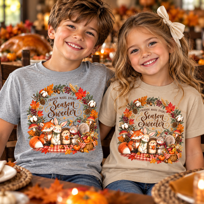 Two children sitting side by side wearing matching graphic t-shirts in a cozy autumn-themed setting. The boy wears a heather gray t-shirt and the girl wears a beige t-shirt, both featuring a colorful circular design with foxes, a bunny, squirrels, pumpkins, and the text 'FINDING MAKE EVERY Season Sweeter'.
