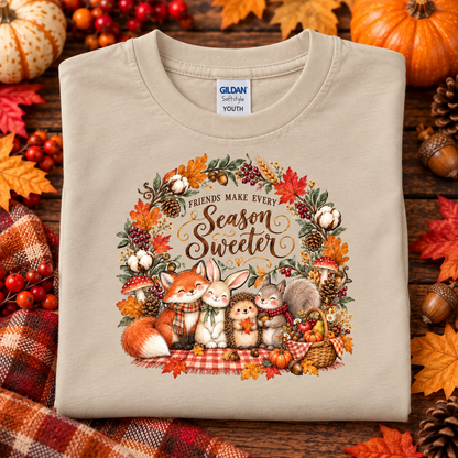 A folded sand Gildan Softstyle Youth t-shirt with a 'Friends Make Every Season Sweeter' autumn graphic, displayed on a rustic wooden background with pumpkins, fall leaves, and a plaid blanket.