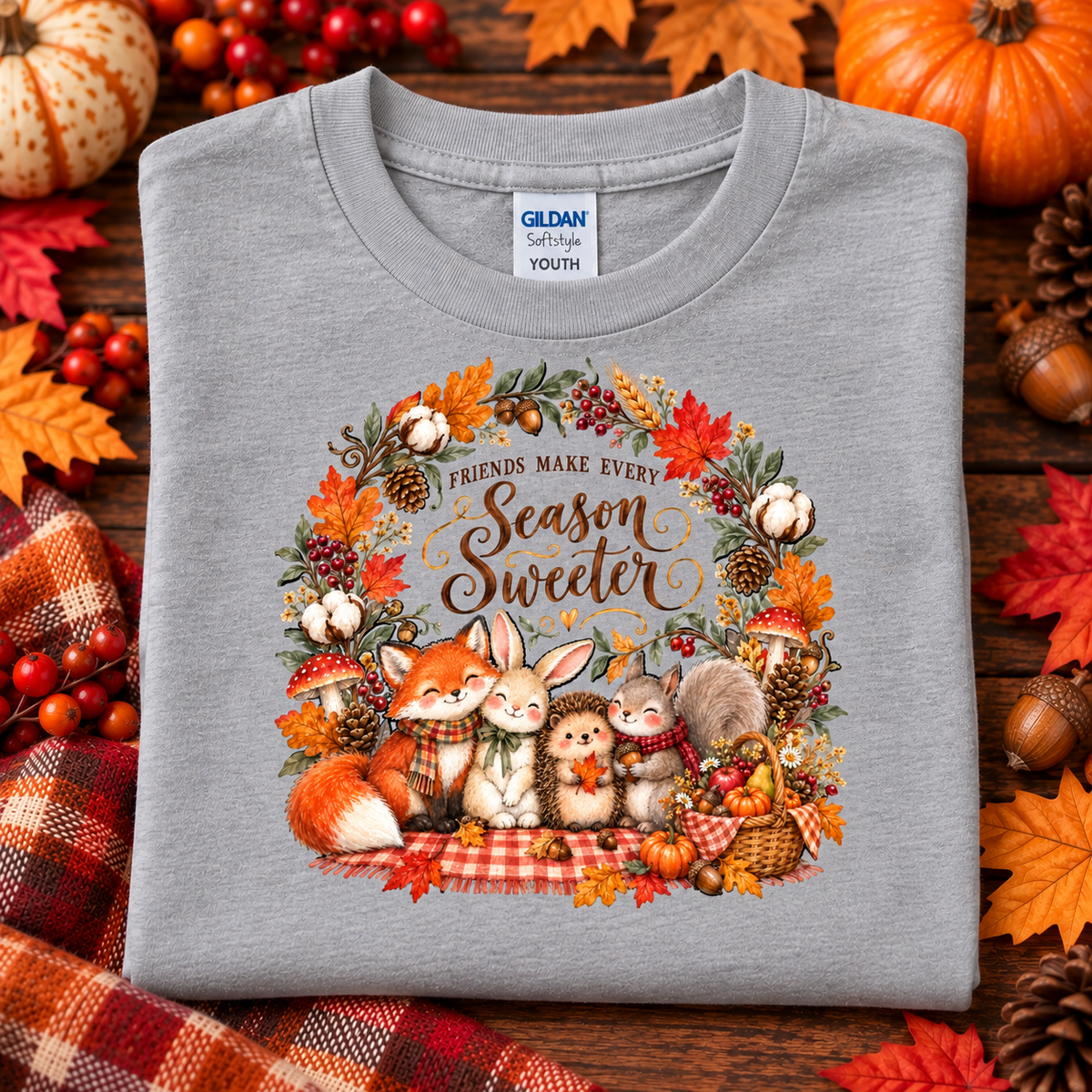 A folded heather gray Gildan Softstyle youth t-shirt with a colorful autumn-themed graphic print, displayed on a rustic wooden background decorated with pumpkins, fall leaves, and a plaid blanket.
