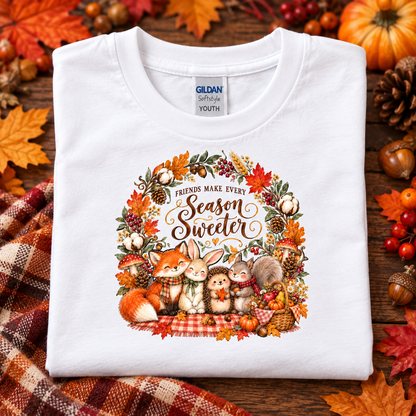 A folded white Gildan Softstyle Youth t-shirt with a colorful autumn-themed graphic print featuring cartoon animals and the text 'Friends Make Every Season Sweeter'. The shirt is displayed on a rustic wooden background surrounded by fall leaves, pumpkins, and a plaid blanket.