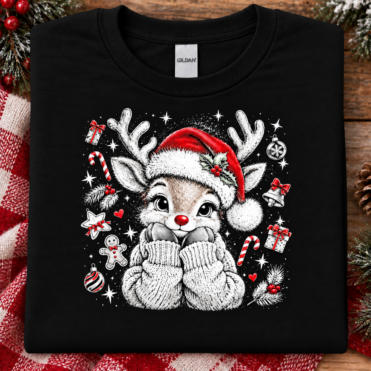 A folded black Gildan t-shirt with a festive Christmas graphic of a cartoon reindeer wearing a Santa hat and white knit mittens, surrounded by candy canes, ornaments, and gifts, displayed on a rustic wooden background with holiday decorations.