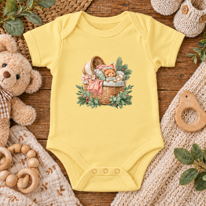 A pale yellow baby bodysuit laid flat on a rustic wooden surface, featuring a central graphic of a wicker basket filled with a teddy bear, pink ribbon, and green foliage. The bodysuit is styled with a plush teddy bear, knit socks, wooden teething rings, and a cream knit blanket.