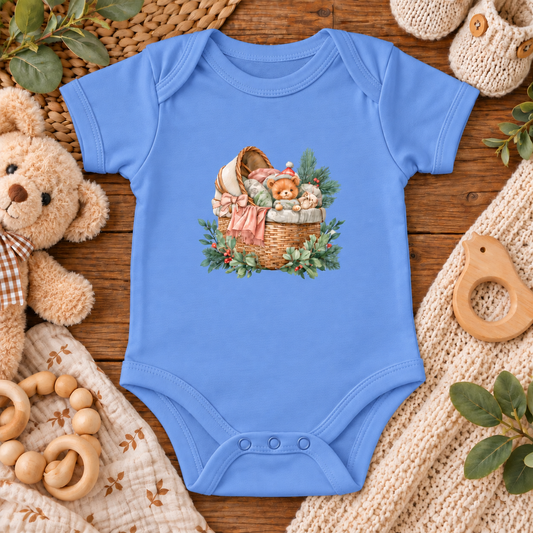 A light blue short-sleeve baby bodysuit laid flat on a rustic wooden surface, featuring a central graphic of a wicker basket filled with teddy bears, a pink bonnet, and green foliage. The bodysuit is styled with baby toys and soft textiles, including a plush teddy bear, wooden rings, a knit blanket, and a crochet teething ring.
