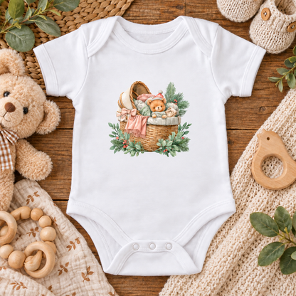 A white short-sleeve baby bodysuit laid flat on a rustic wooden surface, featuring a central illustration of a wicker basket with a teddy bear, pink ribbon, and green foliage. The bodysuit is styled with baby props including a plush teddy bear, wooden rings, a knit blanket, and green leaves.