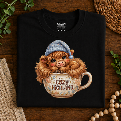 A folded black Gildan t-shirt displayed flat on a rustic wooden surface, featuring a cute graphic of a fluffy Highland cow wearing a blue knit hat and sitting in a floral teacup with the words 'COZY HIGHLAND'.