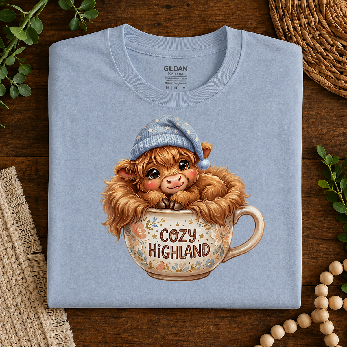 A folded light blue Gildan t-shirt with a cute graphic of a fluffy Highland cow in a blue knit hat sitting in a floral teacup that says 'COZY HIGHLAND', displayed on a dark wood background with decorative greenery and beads.