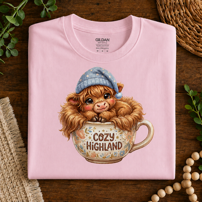 A folded light pink Gildan t-shirt with a cute graphic of a fluffy brown Scottish highland cow wearing a blue knit hat and sitting in a floral mug with the text 'COZY HIGHLAND'. The shirt is styled flat on a dark wood background with decorative greenery, a woven mat, and wooden beads.