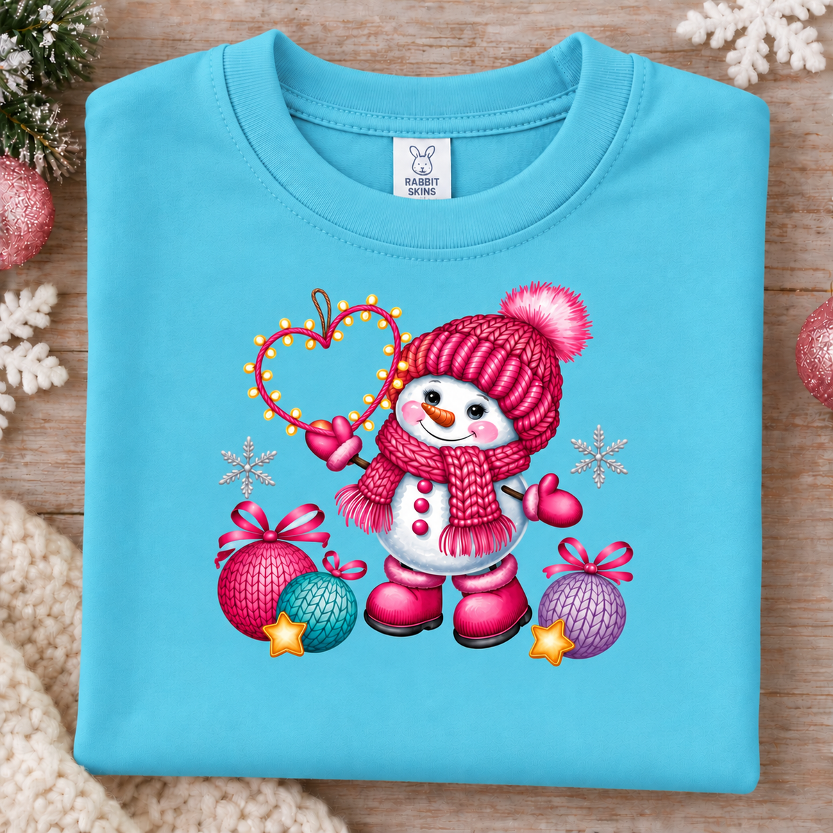 A folded light blue crewneck sweatshirt with a colorful Christmas snowman graphic on the front, displayed on a wooden surface with holiday decorations.