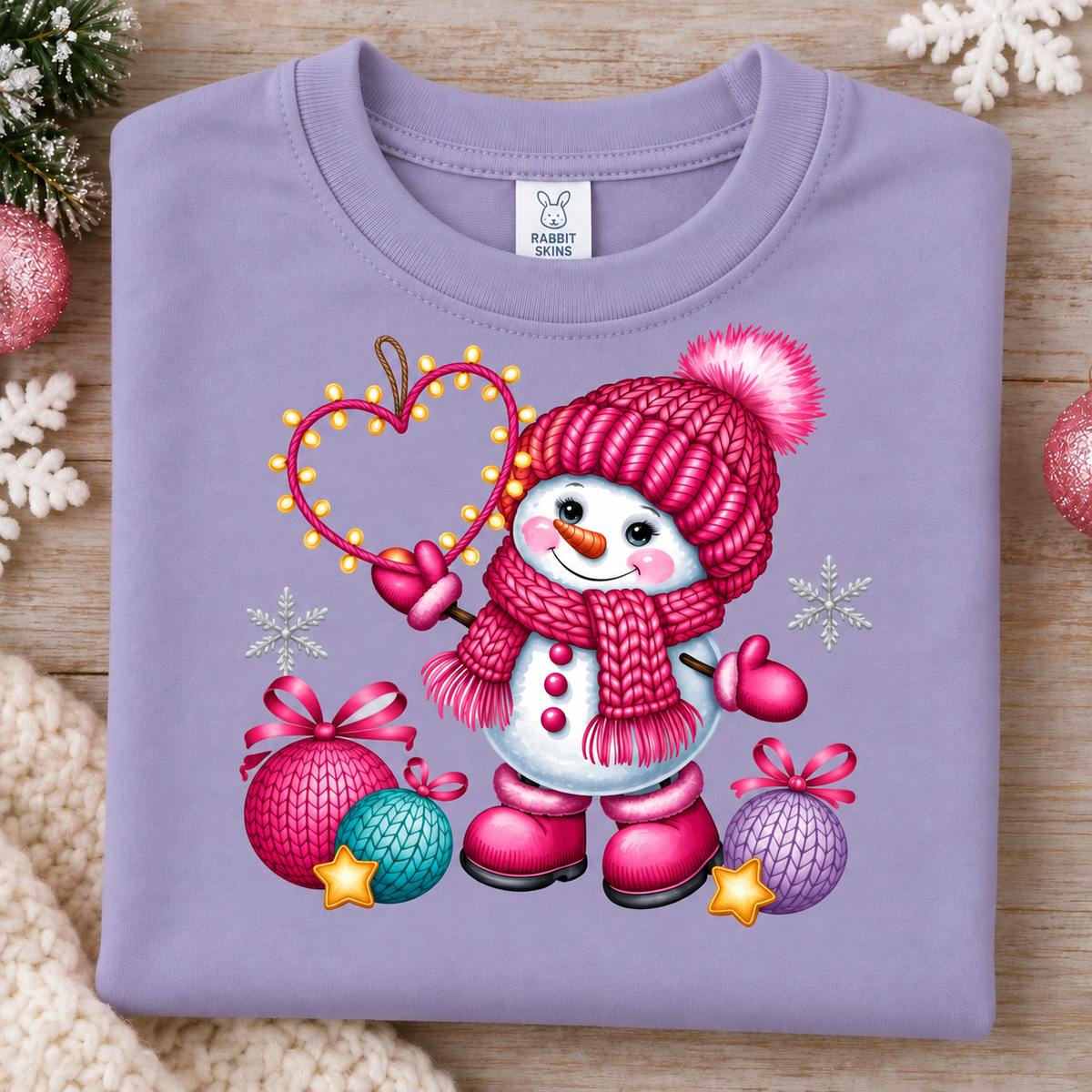 A folded lavender crewneck sweatshirt with a colorful Christmas snowman graphic, displayed on a wooden surface with holiday decorations.