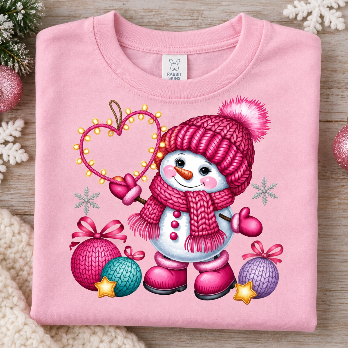 A folded light pink crewneck sweatshirt featuring a colorful graphic of a cartoon snowman wearing a pink knit hat and scarf, surrounded by Christmas ornaments and snowflakes, displayed on a wooden surface with holiday decorations.