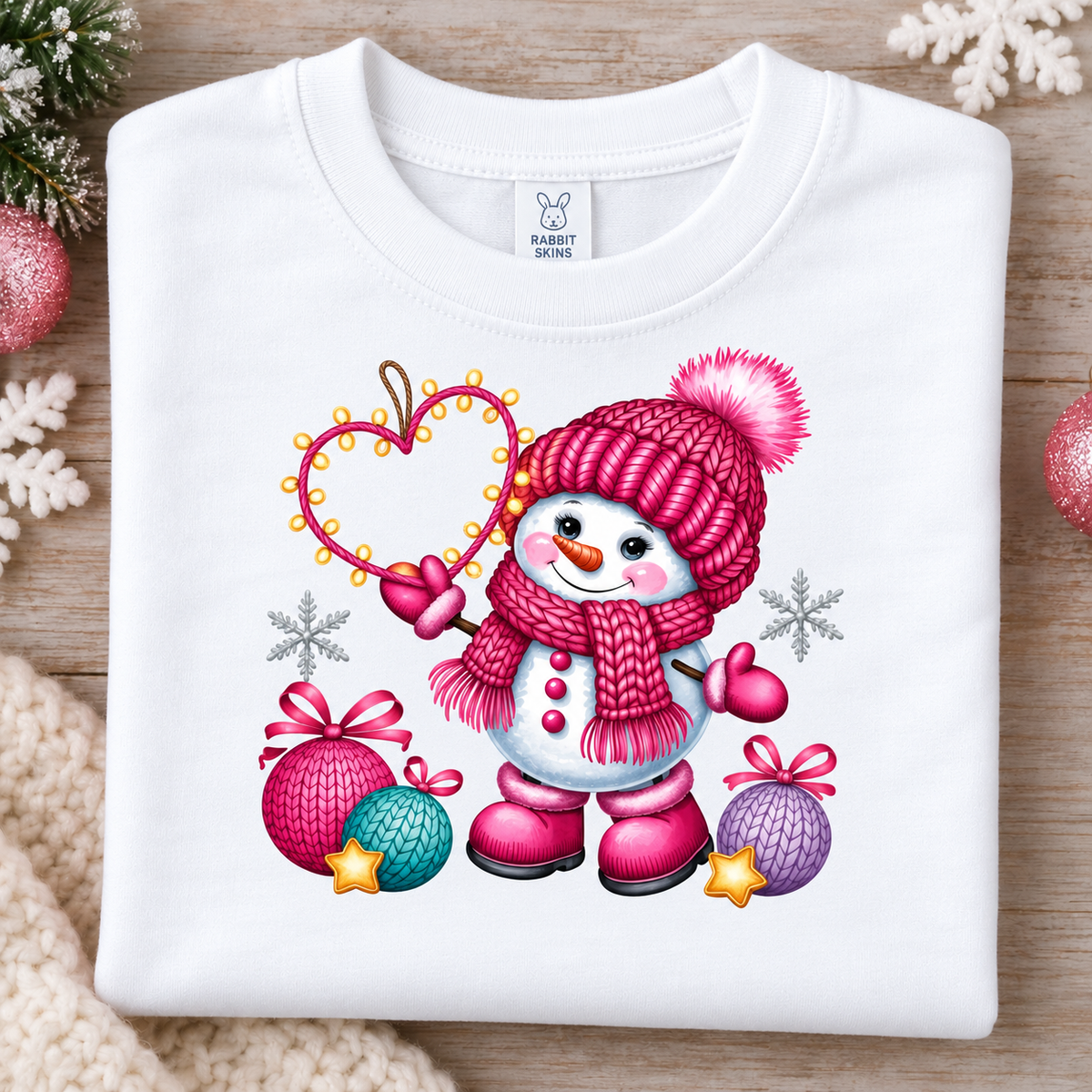 A folded white long-sleeve t-shirt with a colorful Christmas snowman graphic, displayed on a wooden surface with holiday decorations.