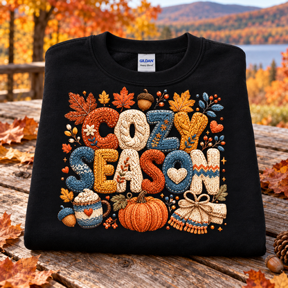 A folded black Gildan sweatshirt with a colorful autumn-themed 'COZY SEASON' graphic, displayed on a rustic wooden table with a blurred fall landscape background.