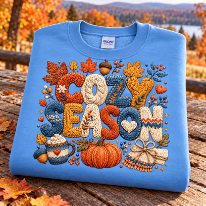 A folded carolina blue crewneck sweatshirt with a colorful autumn-themed 'COZY SEASON' graphic, displayed on a rustic wooden table with a blurred fall landscape background.