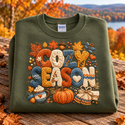 An military green folded crewneck sweatshirt with a large, detailed autumn-themed embroidered graphic reading 'COZY SEASON' surrounded by leaves, pumpkins, and gifts, displayed on a wooden surface with an outdoor fall landscape in the background.