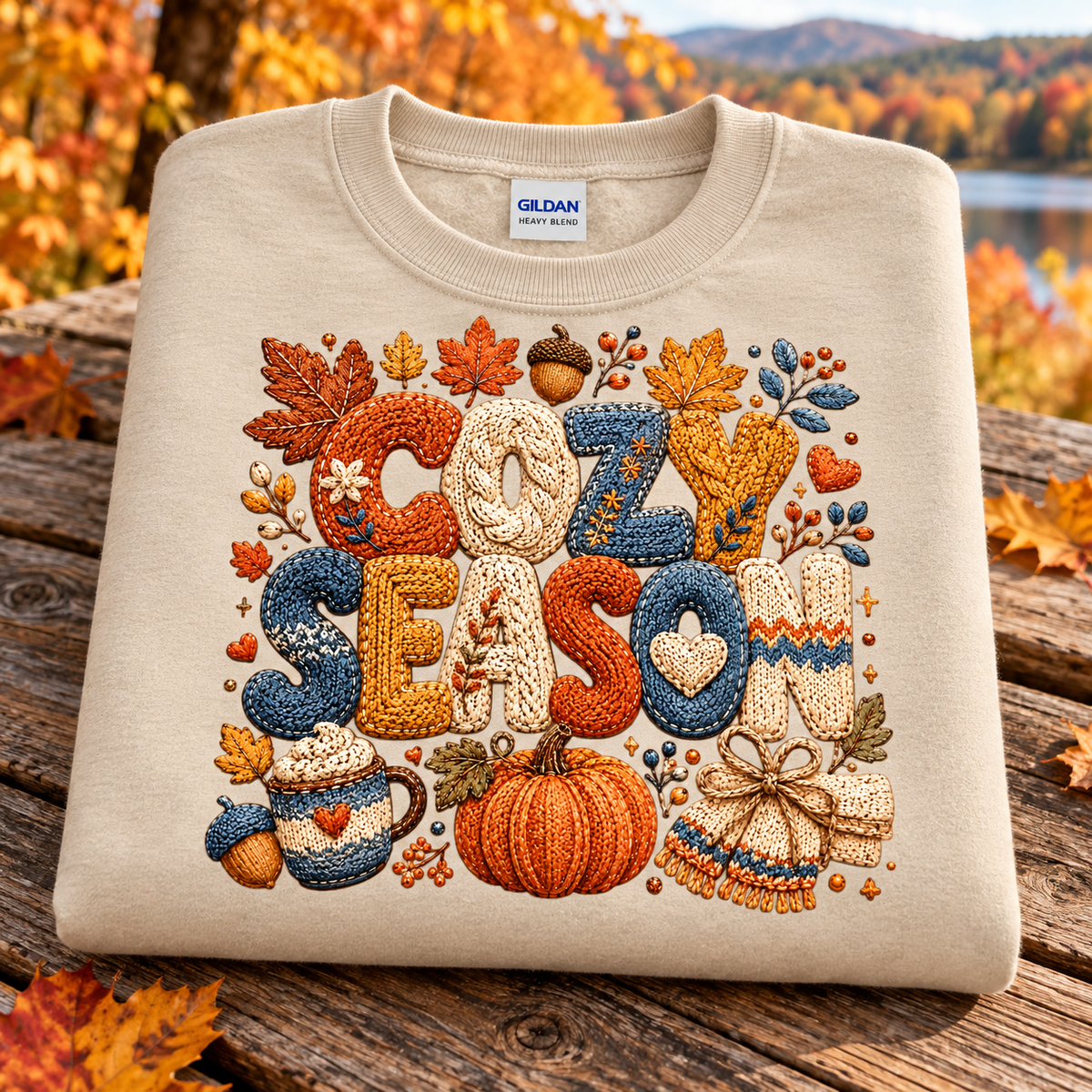 A folded sand colored Gildan sweatshirt with a detailed autumn-themed embroidered design spelling 'COZY SEASON' in warm orange, blue, and cream tones, resting on a rustic wooden surface with a blurred fall landscape background.