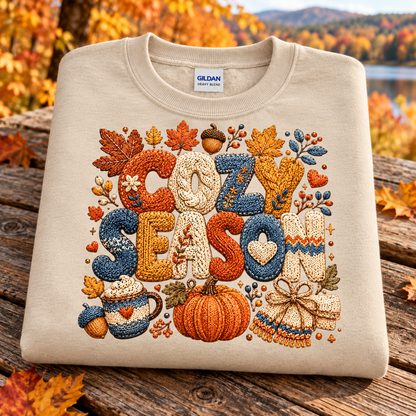 A folded sand colored Gildan sweatshirt with a detailed autumn-themed embroidered design spelling 'COZY SEASON' in warm orange, blue, and cream tones, resting on a rustic wooden surface with a blurred fall landscape background.