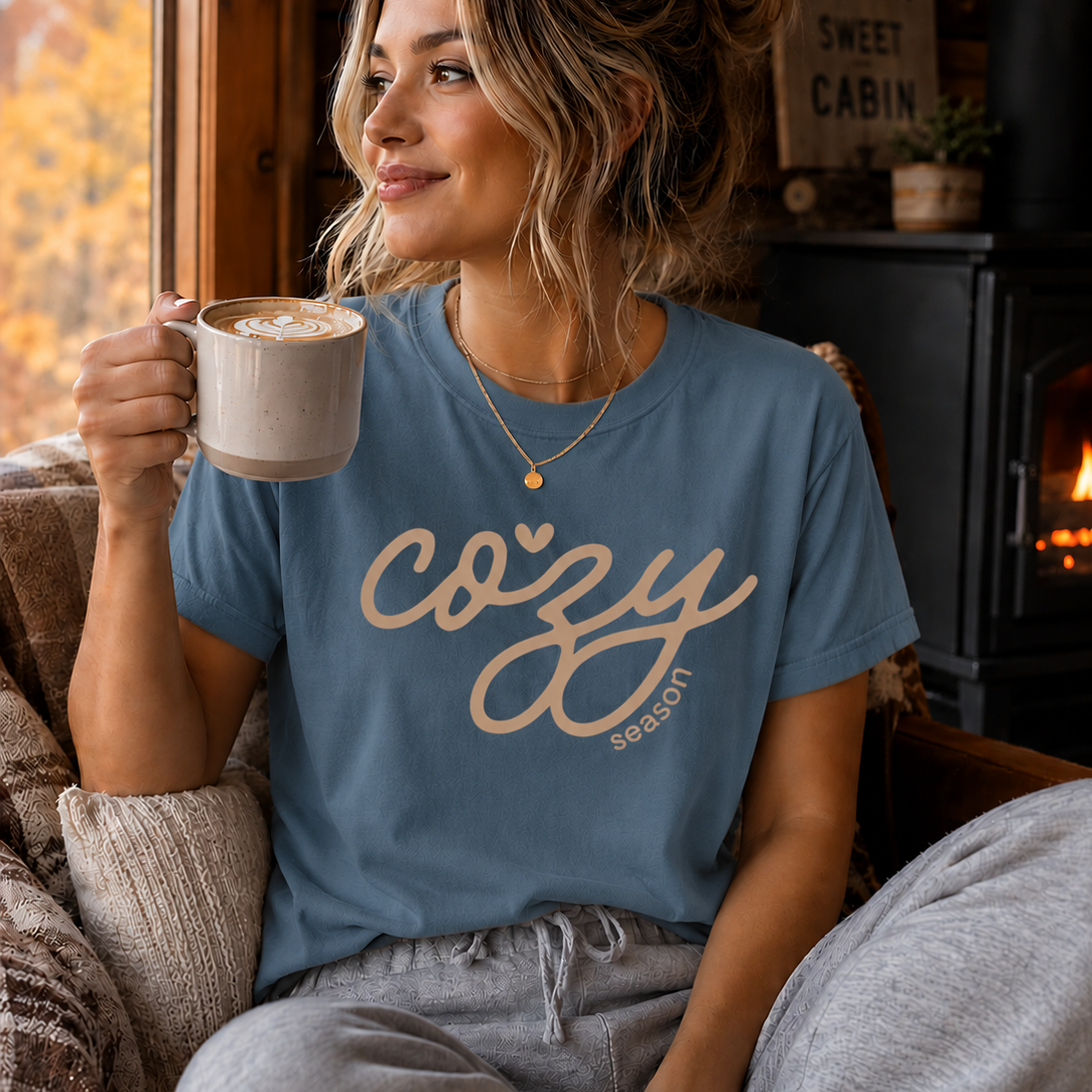 A woman sitting in a cozy room by a window, wearing a ice blue graphic t-shirt with 'cozy season' printed in beige script, holding a mug of coffee. She is wearing gray sweatpants, and a wood-burning stove and fireplace are visible in the background.