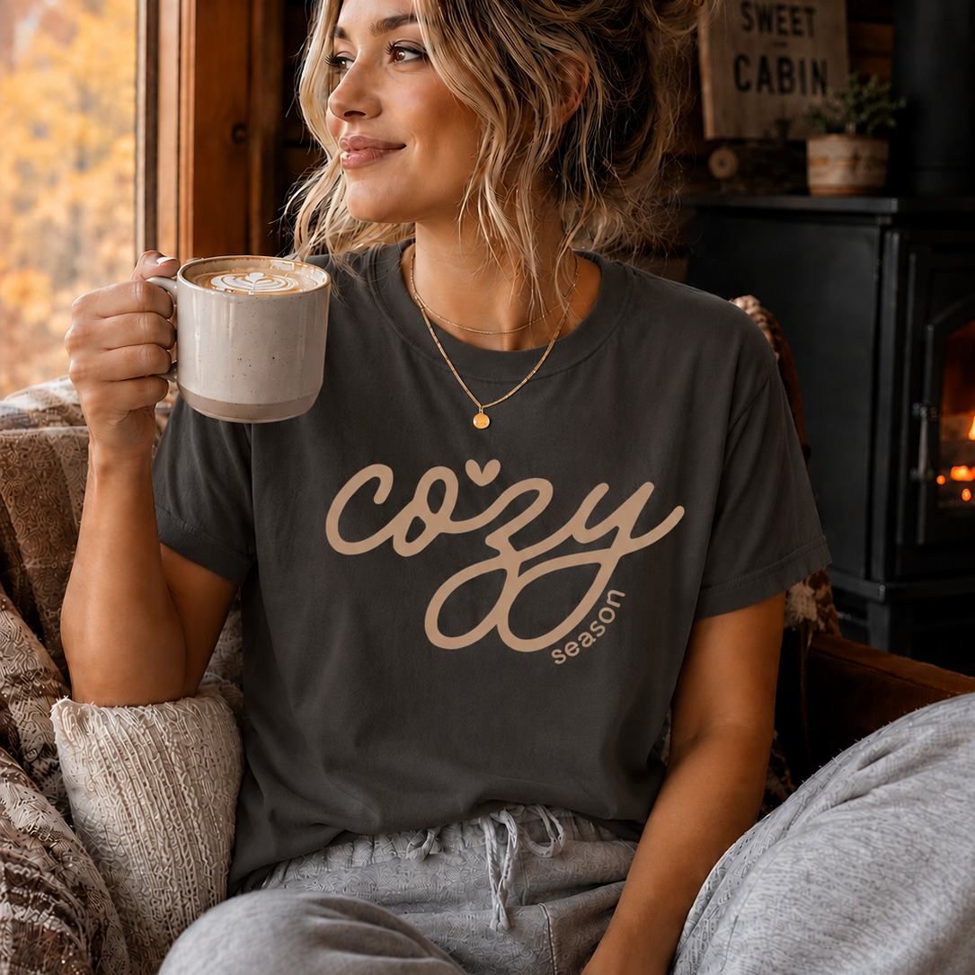 A woman sitting in a cozy cabin setting wearing a pepper colored t-shirt with 'cozy season' printed in pink script, holding a mug and looking out a window.