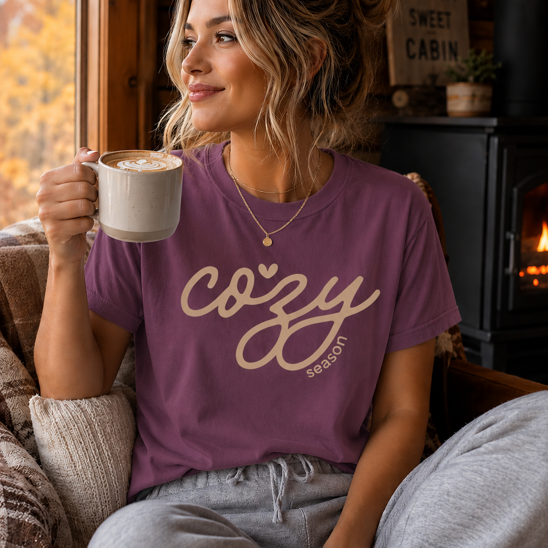 A woman sitting in a cozy cabin setting wearing a berry colored t-shirt with 'cozy season' printed in pink script, holding a mug of coffee.