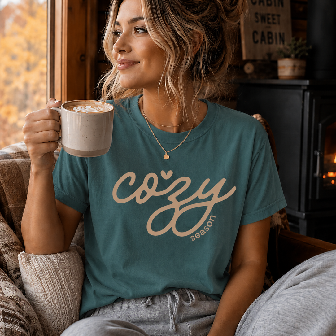 A woman sitting in a cozy cabin setting wearing a seafoam t-shirt with 'cozy season' printed in beige script, holding a mug. The background shows a warm, rustic interior with a fireplace and autumn trees outside the window.
