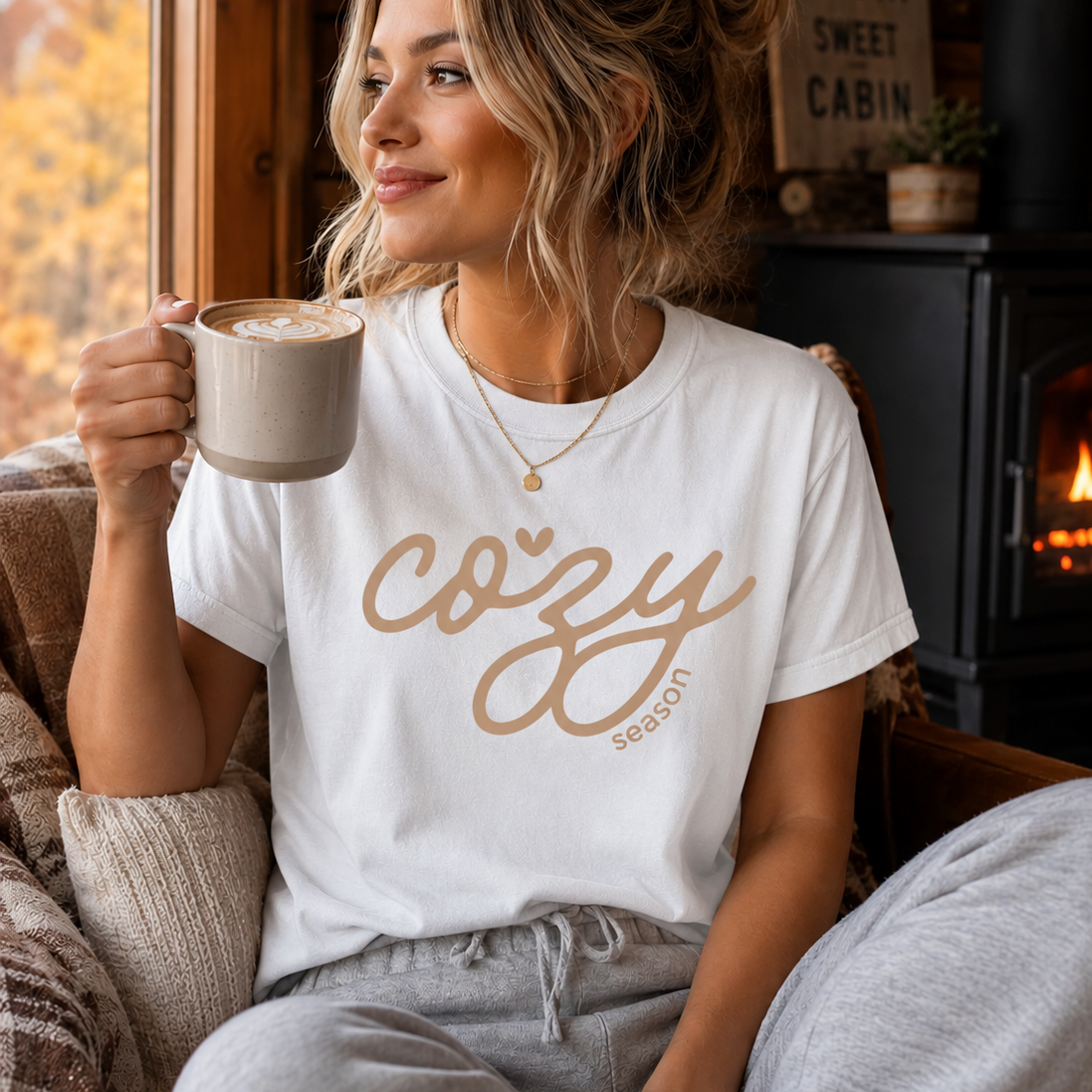 A woman sitting in a cozy cabin setting wearing a white graphic t-shirt that reads 'cozy season' in a pink script, holding a mug of coffee.