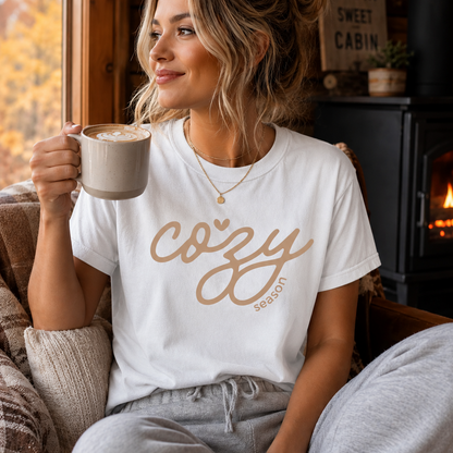 A woman sitting in a cozy cabin setting wearing a white graphic t-shirt that reads 'cozy season' in a pink script, holding a mug of coffee.