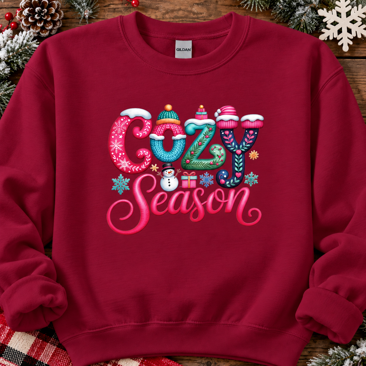 A cardinal red crewneck sweatshirt featuring a colorful 'Cozy Season' graphic with winter-themed lettering, styled on a rustic wooden background with pinecones and holiday decorations.